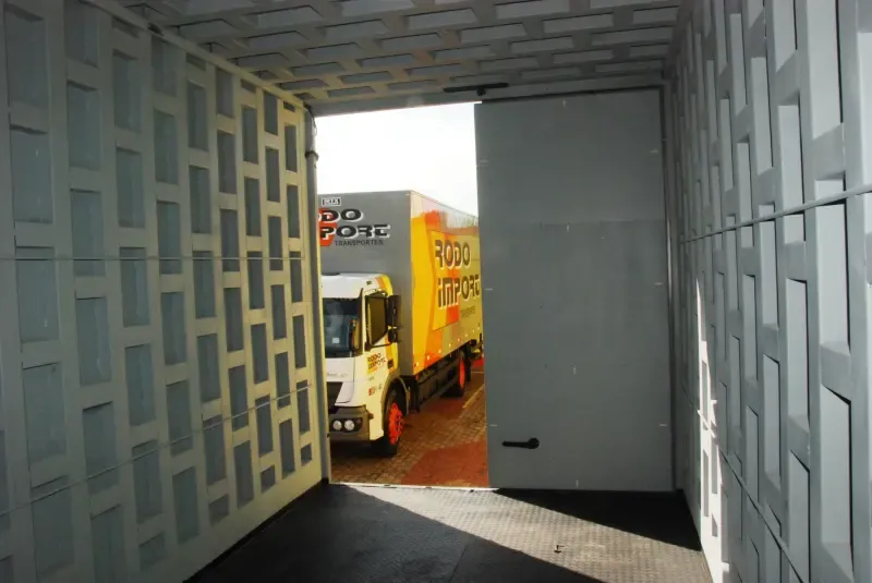Vista de dentro de um baú com porta-cofre aberta, com caminhões da Rodo Import ao fundo